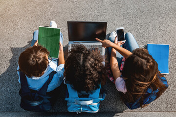 Happy group of preteen school kids children elementary middle classmates sitting on the stairs and using digital devices laptop smartphone and notebook doing homework outdoors