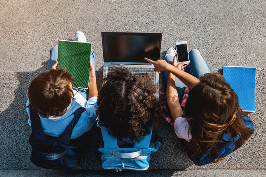 Happy group of preteen school kids children elementary middle classmates sitting on the stairs and using digital devices laptop smartphone and notebook doing homework outdoors