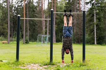 An athletic man in gravity boots hangs on a horizontal bar outdoors on a summer day.