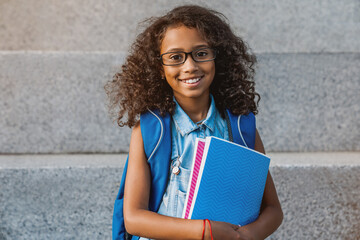 Portrait of happy african young preteen girl elementary middle school pupil in eyeglasses with backpack holding notebook books outside the primary school. Education concept