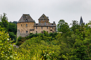 Naklejka premium Reinhardstein Castle seen from a viewing point, towers and stone walls with multiple windows, Warche Valley mountain surrounded by green leafy trees, cloudy summer day in Waimes, Belgium