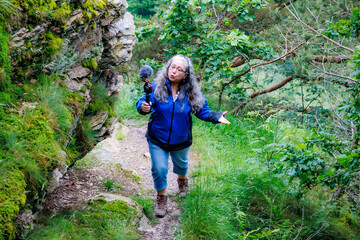 Senior adult woman walking on trail and making videos around ruined castle Logne, talking to camera, rocky wall, wild vegetation and trees in background, blue jacket, cloudy day in Ferrieres, Belgium