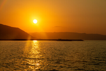 A serene sunset over the mountains reflects golden hues on the water. The vibrant orange and yellow sky creates a peaceful, captivating scene as the sun dips below the horizon.