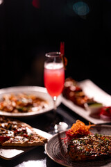 Red cocktail drink in glass on dark background with foods on table. Pizza, Pasta and Kebab platter with Cocktail.