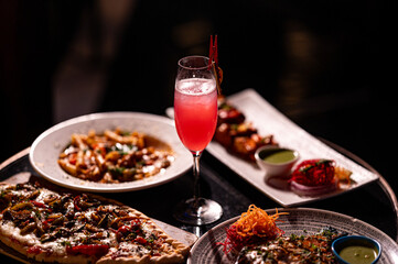 Red cocktail drink in glass on dark background with foods on table. Pizza, Pasta and Kebab platter with Cocktail.