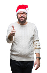 Young caucasian man wearing christmas hat and glasses over isolated background doing happy thumbs up gesture with hand. Approving expression looking at the camera with showing success.