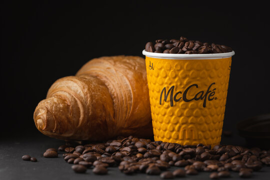 KYIV, UKRAINE - January 06: Close up of yellow McDonald's paper cup of coffee with a fresh croissant in a popular fast food restaurant. McCafe cup with flying coffee beans on the black background