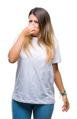 Young beautiful woman casual white t-shirt over isolated background smelling something stinky and disgusting, intolerable smell, holding breath with fingers on nose. Bad smells concept.