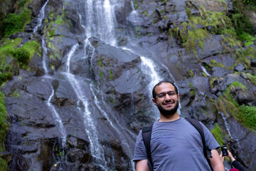 A smiling man stands near a cascading waterfall on wet, dark rocks, wearing a backpack and outdoor gear, enjoying a nature hike