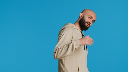 Middle eastern guy asking people to come over, posing over studio background. Arab person invites someone to join him, welcome to come closer on camera, casual invitation. Camera 1.