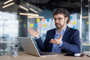 Man wearing suit eyeglasses engaged video conference call using laptop and headphones in modern office setting, discussing work with expressive hand gestures, technology, communication, remote work