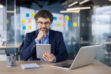 Fototapeta premium Confident businessman in suit using digital tablet while working in modern office. Professional workspace includes laptop, notebook, and glass walls with sticky notes