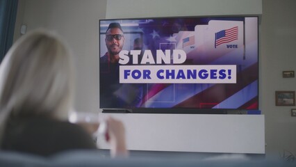 Adult woman sits on sofa at home with cup of coffee and watches political advertising or campaign about United States presidential elections on big TV screen. TV live broadcast. National Election Day.