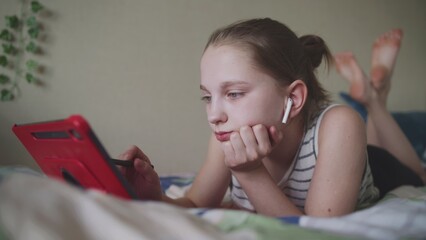 Young girl lies on bed at home, surfs the Internet or scrolls social networks using tablet computer and digital pencil. Caucasian teenager spends leisure time in comfort bedroom. Lifestyle concept. © Framestock