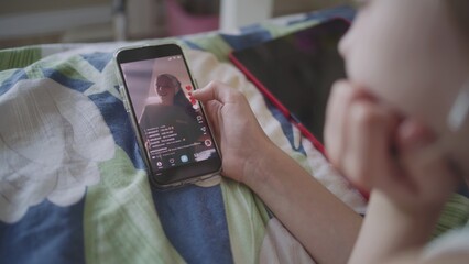 Teenager lies on bed, watches video content in social network using mobile phone. Caucasian girl...