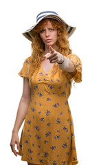 Young redhead woman wearing summer hat and dress pointing with finger to the camera and to you, hand sign, positive and confident gesture from the front