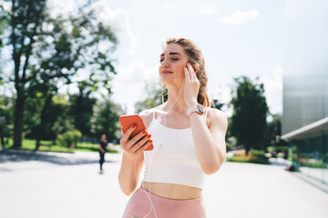 Young Caucasian woman savoring music on her smartphone while outdoors on a bright summer day. Clad in sporty clothes, wearing earphones and a smartwatch, exuding relaxation and joy in