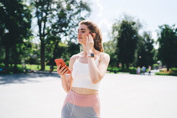 Young Caucasian woman enjoying music on smartphone while standing outdoors in sunny park. Wearing sporty attire, with earphones and smartwatch, looking relaxed and content, surrounded by greenery