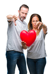 Middle age hispanic casual couple in love holding red heart over isolated background with angry face, negative sign showing dislike with thumbs down, rejection concept