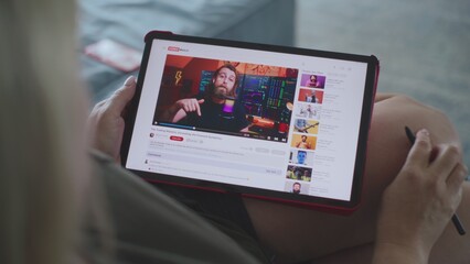 Caucasian woman watches educational video of blogger about stock trading using tablet computer while lying on sofa. Tablet screen shows video hosting website interface with video content. Close Up. © Framestock