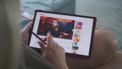 Caucasian woman watches educational video of blogger about stock trading using tablet computer while lying on sofa. Tablet screen shows video hosting website interface with video content. Close Up.