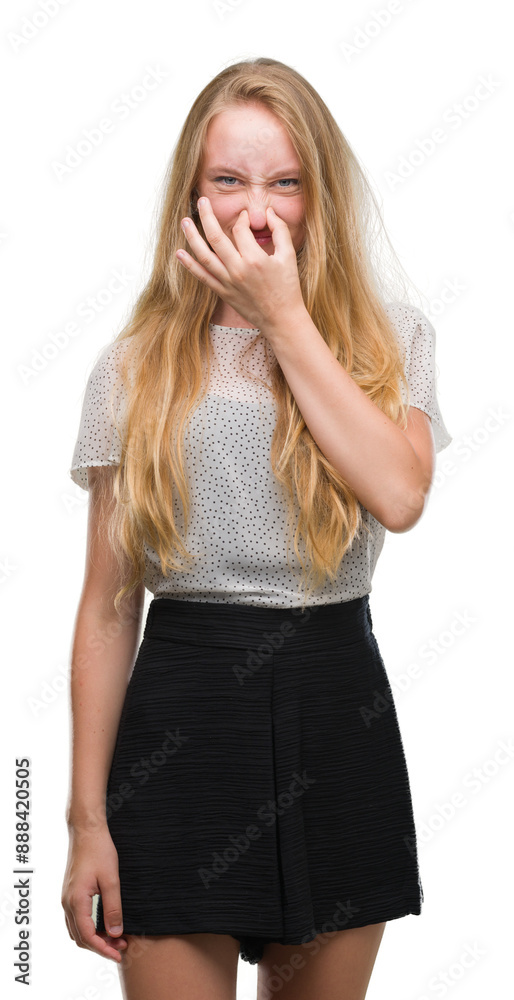 Wall mural Blonde teenager woman wearing moles shirt smelling something stinky and disgusting, intolerable smell, holding breath with fingers on nose. Bad smells concept.