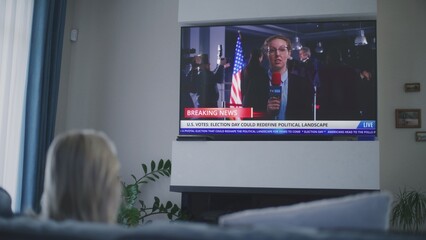 Woman rests on couch in living room, watches live broadcast on TV. African American female correspondent reports breaking news live on Election Day in the USA. Civic duty and patriotism. High angle.