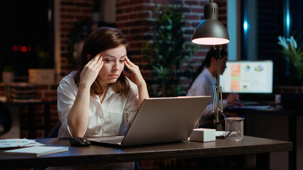 Tired employee struggling to remain focused at workspace desk, feeling overworked and waiting to go home. Bored businesswoman sighing in office while working overnight, camera B