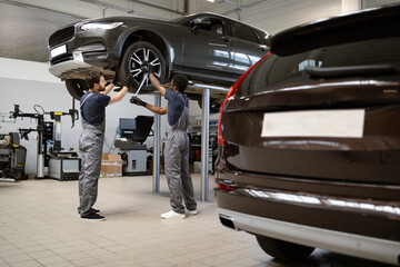Two mechanics in uniform repairing car on lift in modern auto repair shop. Professional technicians...