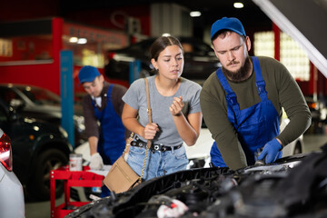 Young guy mechanic advises young woman client on repairing under hood of car in car service station