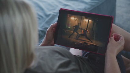 Close up of woman watching action movie using tablet computer while lying on sofa. Caucasian woman relaxes at home on the weekend. Digital tablet screen with displayed thriller film. Lifestyle concept