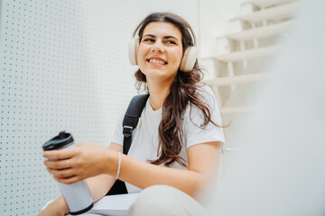One young caucasian woman student sitting on stairs near university listening to music and drinking coffee	