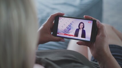 Close up static shot of adult woman watching online TV breaking news broadcast about elections in the United States of America using smartphone while lying on sofa at home. Election Day in USA.