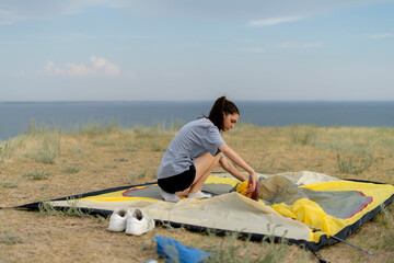 woman wearing a grey shirt and black shorts squats on a beach cliff while setting up a yellow tent