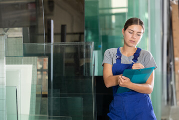 Young female technician in blue overalls carefully inspecting glass panels, focused on quality check, and writing notes on clipboard against blurred workshop background