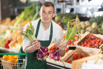 Sales man works in vegetable store bio supermarket and goes through goods. He holds onion in hands, examines it, checks quality