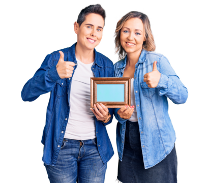 Couple of women holding empty frame doing happy thumbs up gesture with hand. approving expression looking at the camera showing success.