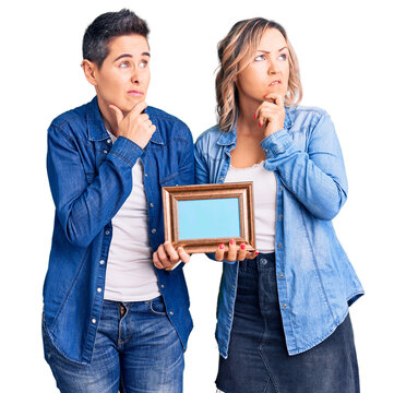Couple of women holding empty frame thinking worried about a question, concerned and nervous with hand on chin