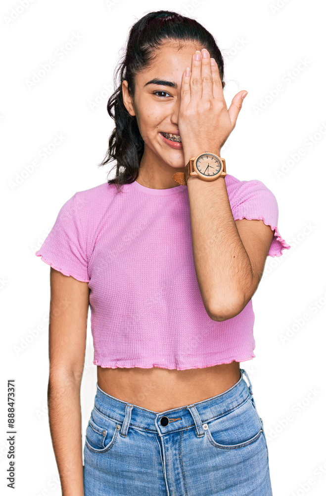 Wall mural Hispanic teenager girl with dental braces wearing casual clothes covering one eye with hand, confident smile on face and surprise emotion.