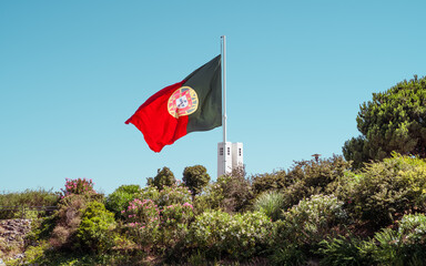 The national flag of Portugal waves prominently on a tall flagpole, set against a clear blue sky. The flag is surrounded by lush greenery, including various trees and shrubs in a defocused foreground