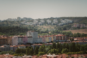 Obraz premium A suburban area near Lisbon featuring colorful residential buildings in the foreground and densely packed apartment blocks on a hillside in the background, surrounded by lush greenery and a clear sky