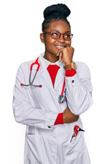 Young african american woman wearing doctor uniform and stethoscope with hand on chin thinking about question, pensive expression. smiling and thoughtful face. doubt concept.