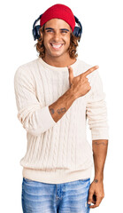 Young hispanic man listening to music using headphones cheerful with a smile of face pointing with hand and finger up to the side with happy and natural expression on face