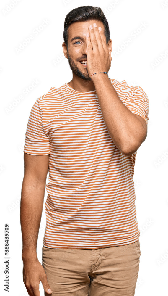 Sticker Handsome hispanic man wearing casual clothes covering one eye with hand, confident smile on face and surprise emotion.