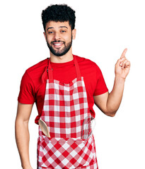 Young arab man with beard wearing cook apron with a big smile on face, pointing with hand and finger to the side looking at the camera.