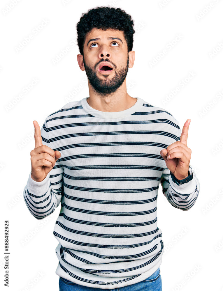 Canvas Prints Young arab man with beard wearing casual striped sweater amazed and surprised looking up and pointing with fingers and raised arms.