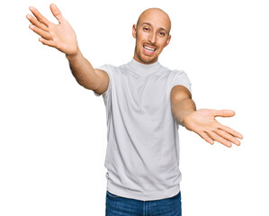 Bald man with beard wearing casual white t shirt looking at the camera smiling with open arms for hug. cheerful expression embracing happiness.