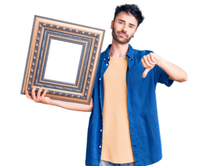 Young hispanic man holding empty frame with angry face, negative sign showing dislike with thumbs down, rejection concept