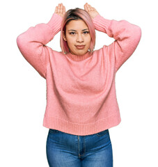 Hispanic woman with pink hair wearing casual winter sweater doing bunny ears gesture with hands palms looking cynical and skeptical. easter rabbit concept.