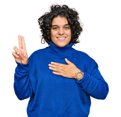 Young hispanic woman with curly hair wearing turtleneck sweater smiling swearing with hand on chest...